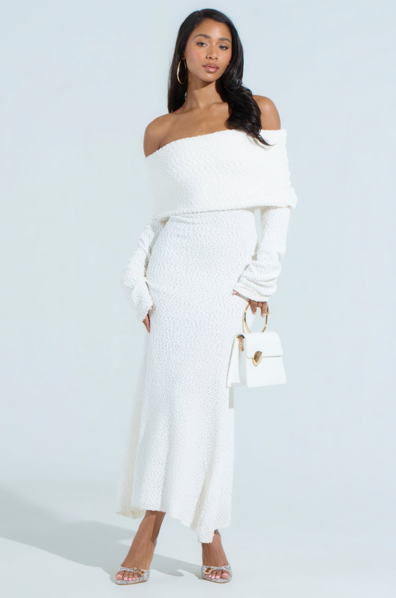 A woman stands against a light backdrop wearing the CANCELLED PLANS KNIT MIDI DRESS, which is off-the-shoulder and curve-skimming in white. She accessorizes with a small white handbag, hoop earrings, and strappy heels, as her long hair cascades over one shoulder.