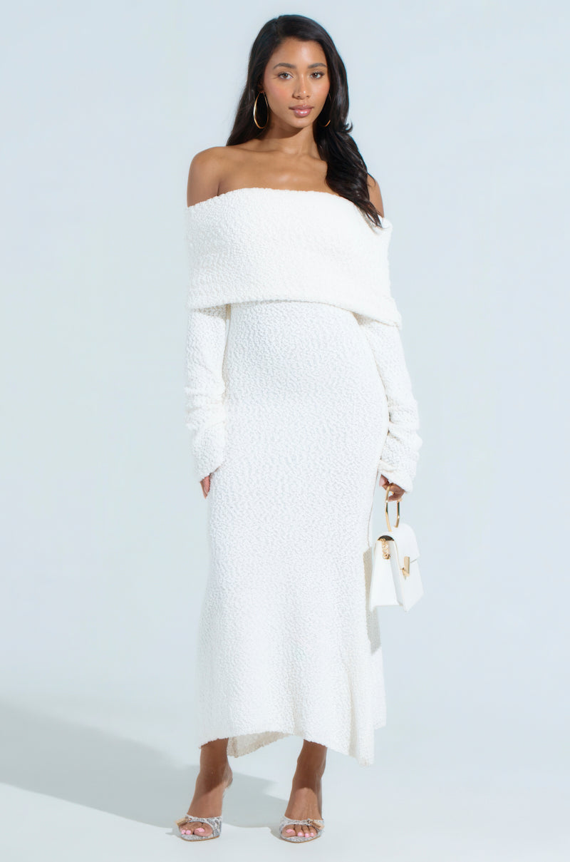 A woman wearing the CANCELLED PLANS KNIT MIDI DRESS with a curve-skimming fit holds a small white handbag. She's adorned with hoop earrings and transparent heels, set against a plain white background.