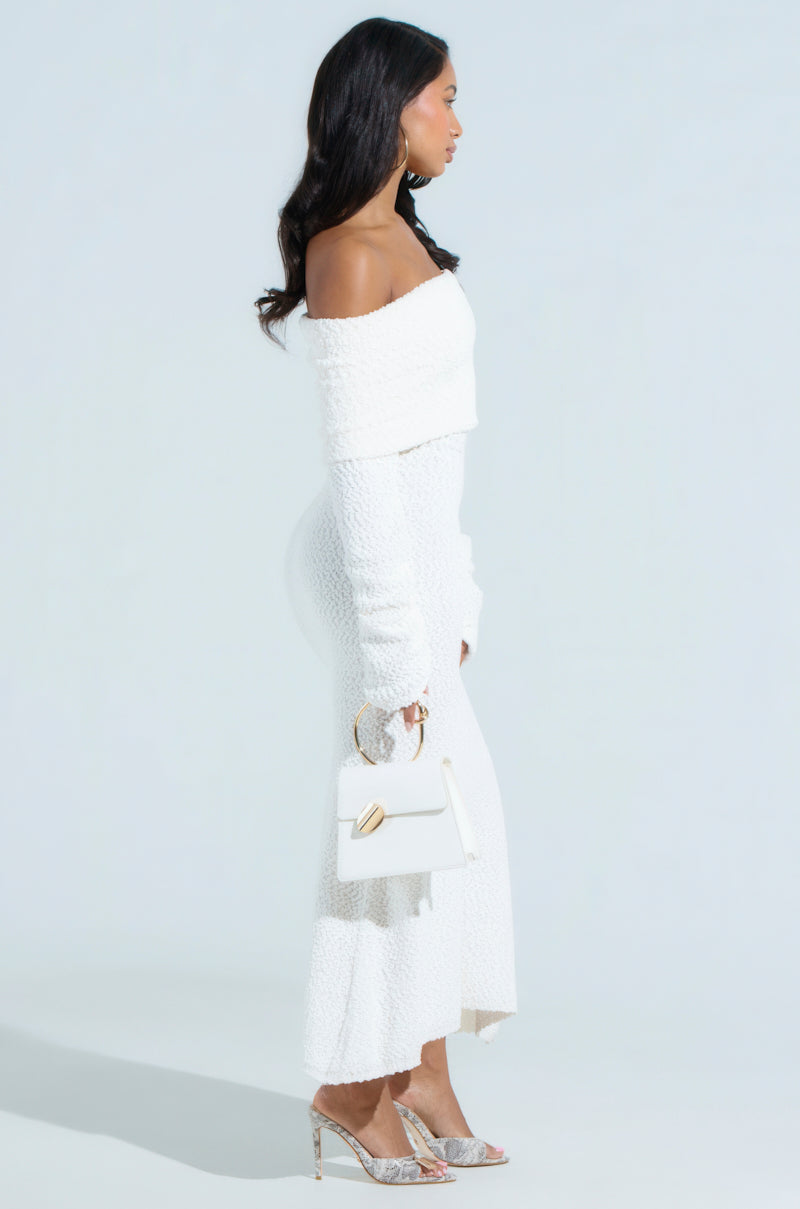 A woman in a white CANCELLED PLANS KNIT MIDI DRESS with an elegant convertible off-the-shoulder design stands in profile against a light backdrop. She holds a small white handbag, wears silver high-heeled sandals, and her long dark hair cascades down her back.