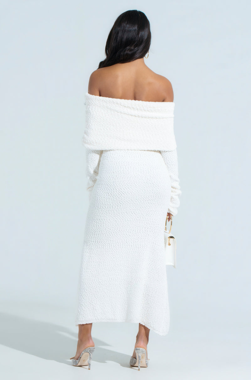A woman with long dark hair models the CANCELLED PLANS KNIT MIDI DRESS, styled in an off-the-shoulder, curve-skimming fit. She's turned away, holding a small white handbag and wearing strappy heeled sandals against a plain, light-colored background.