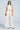 A woman stands against a plain light background wearing the BLAKE SATIN WIDE LEG PANT and a sleeveless, high-neck white satin top. She accessorizes with a gold clutch and strappy heeled sandals.