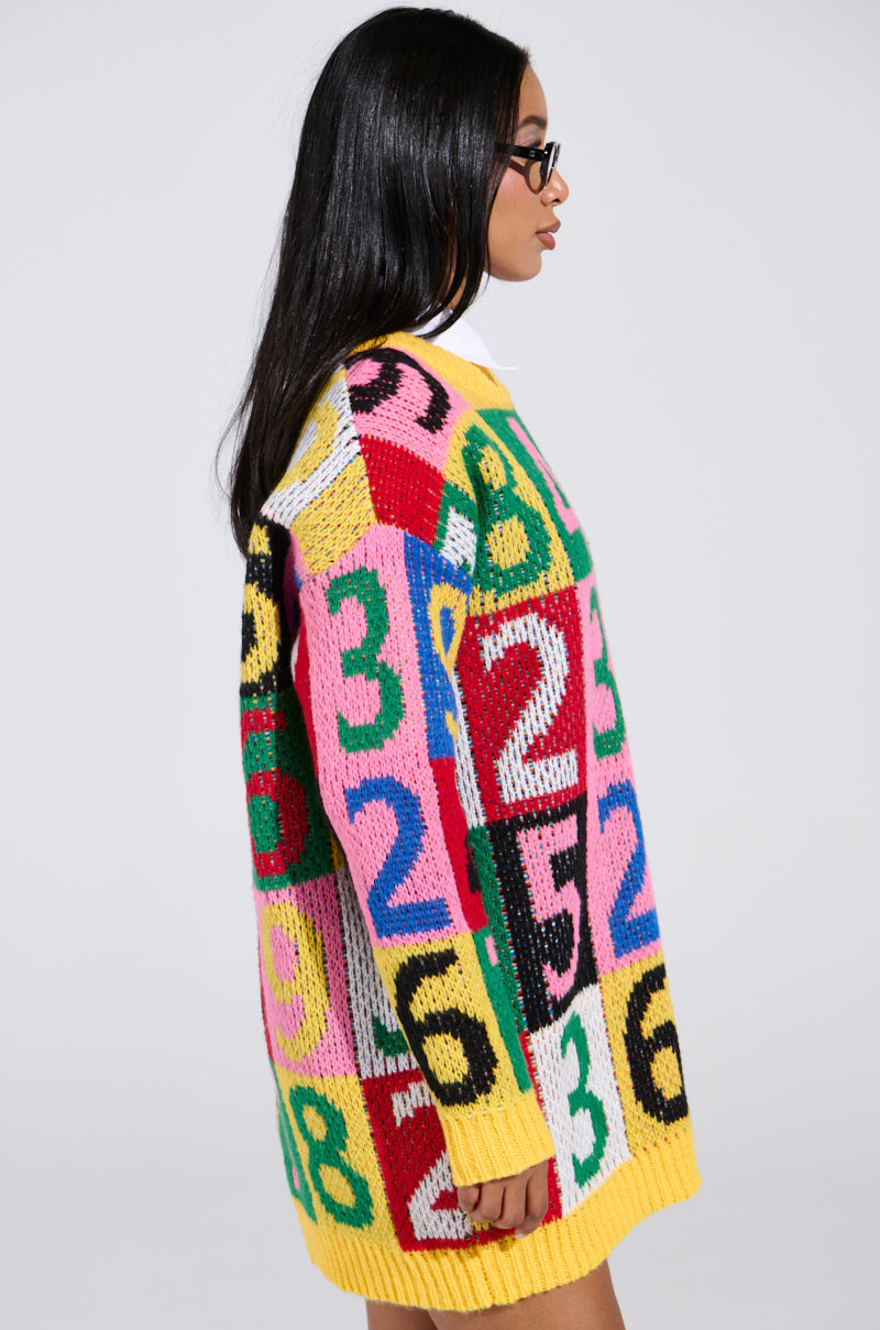 A person with long dark hair and glasses is wearing the QUICK ONE TWO THREE SWEATER DRESS, showcasing a color block design with large numbers in pink, yellow, and black. The plain neutral background highlights the vibrant pattern of this cozy knit fabric.