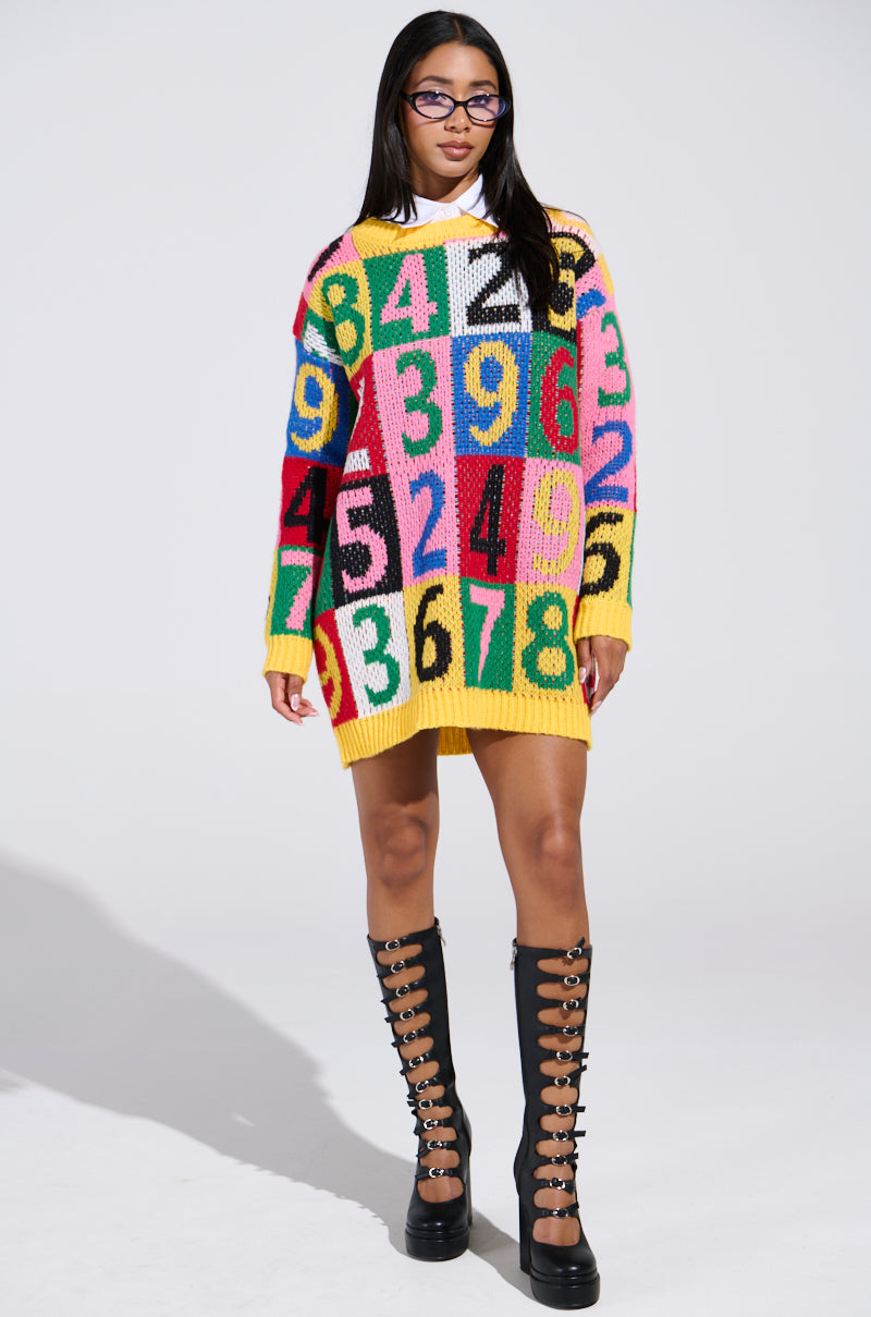 Against a plain background, a woman with long dark hair and glasses wears the QUICK ONE TWO THREE SWEATER DRESS, featuring large numbers and a vibrant color block design. She pairs it with black knee-high boots adorned with multiple buckles.