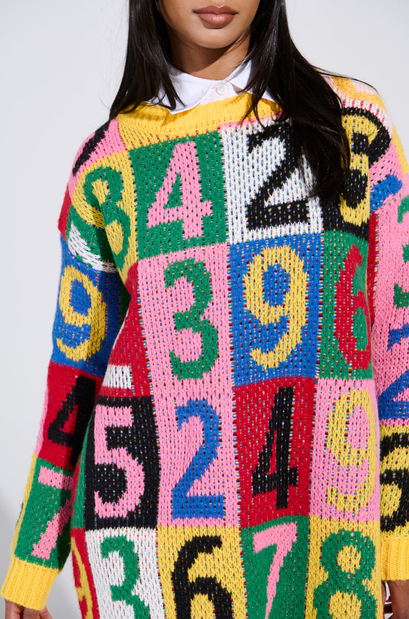 A person dons the QUICK ONE TWO THREE SWEATER DRESS with its cozy knit fabric, large colorful numbers in pink, blue, green, and yellow in a vibrant color block design. Long dark hair frames their face, highlighting a crisp white shirt collar.