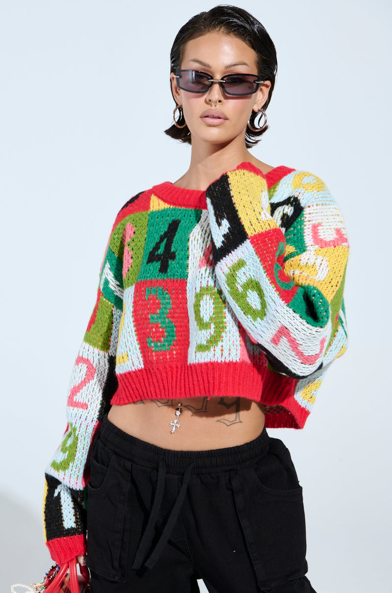 A person shows off the colorful, oversized TOO MANY NUMBERS SWEATER featuring a color-block design with numbers and patterns, paired with black pants and sunglasses. They have short dark hair, hoop earrings, and a belly chain against a plain light blue backdrop.