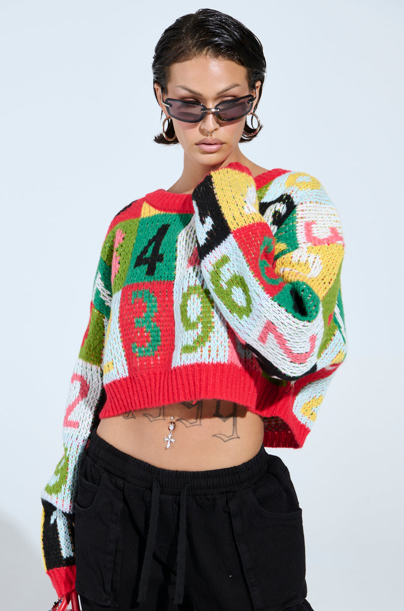 Against a light background, someone in a TOO MANY NUMBERS SWEATER with a colorful color-block design and numbers poses wearing sunglasses. They have short, slicked-back hair, dark pants, and tattoos visible on their midriff below the cropped knit.
