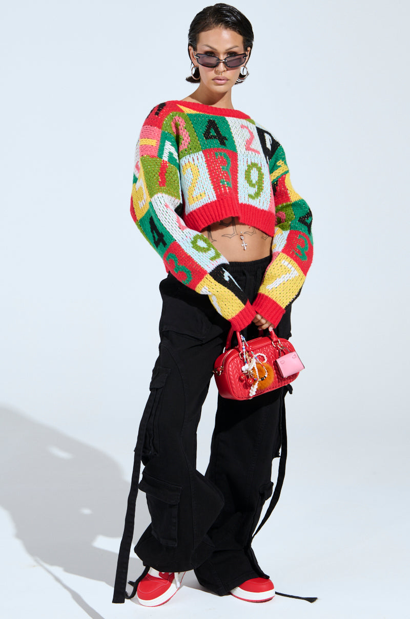 A person poses confidently in the TOO MANY NUMBERS SWEATER, a colorful color-block design cropped sweater with numbers. They wear black cargo pants with straps, sunglasses, and red sneakers, holding a shiny red bag with gold details against a plain white background.
