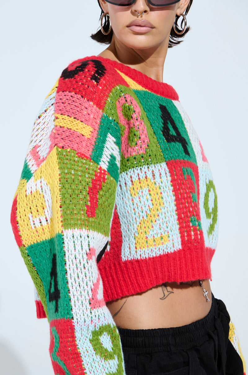 The person showcases a TOO MANY NUMBERS SWEATER with a vibrant color-block design featuring bold red, green, yellow, and pink numbers on a stretchy knit. They're accessorized with sunglasses, hoop earrings, and a nose ring while black pants are visible at the bottom of the frame.
