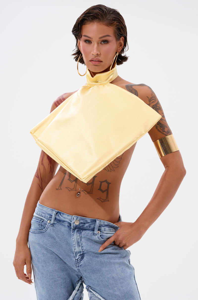 A person with short hair and tattoos wears blue jeans, a shiny yellow halter top, the KATE OVERSIZED NECKLACE, gold hoop earrings, and a wide gold cuff, posing against a plain light background.