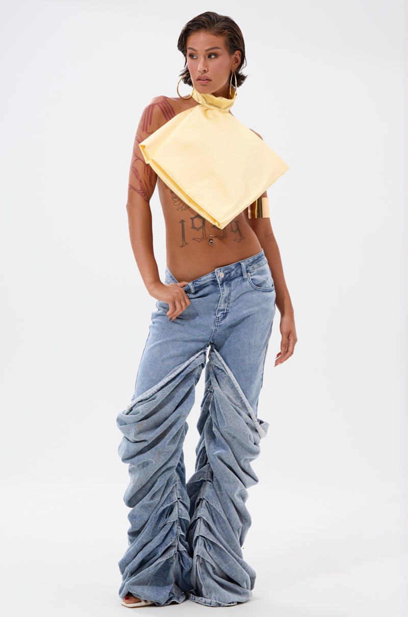 A person poses confidently in ruched, wide-leg blue jeans and a sheer brown top, styled with the KATE OVERSIZED NECKLACE—an unmistakable statement piece—set against a plain white background.
