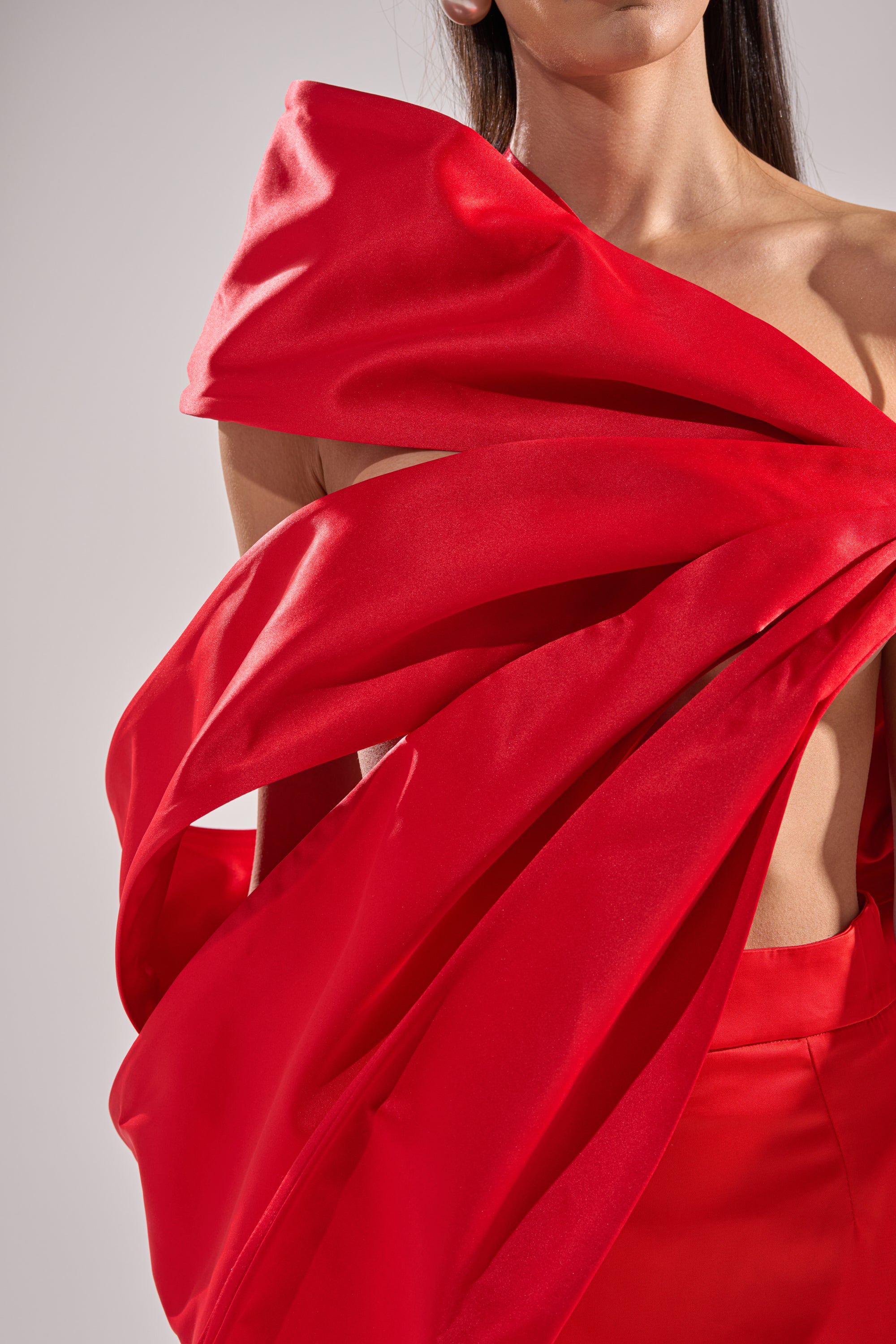 A person wears the IM THE GIFT BODY ACCESSORY—a bold, asymmetrical red satin top with an oversized sculptural bow—paired with matching red pants, set against a neutral background.