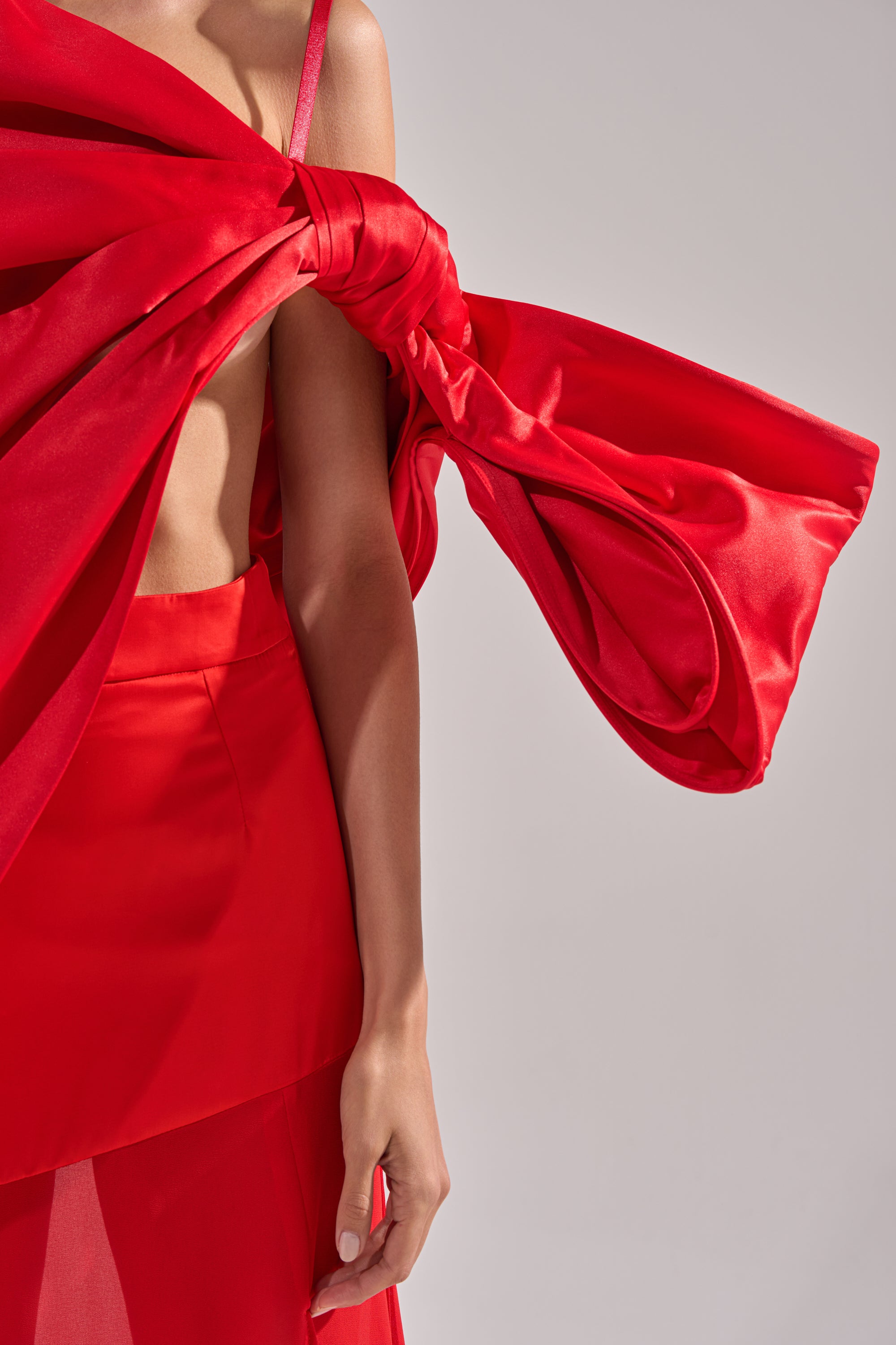 A person wears the IM THE GIFT BODY ACCESSORY, a bold red satin oversized bow draped across the chest and shoulder, partially revealing their torso and arm, against a neutral background.