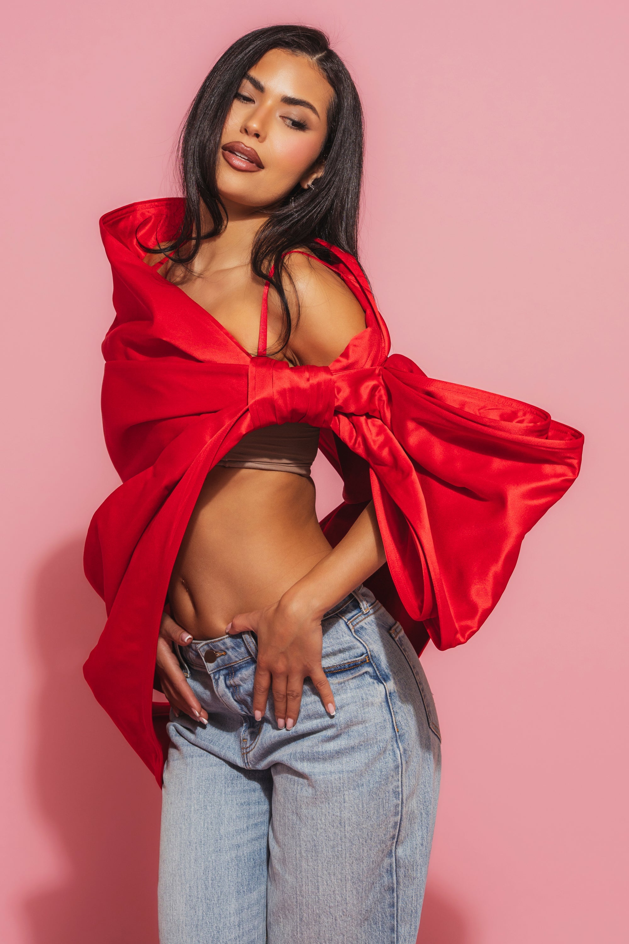 A woman with long dark hair poses confidently against a pink background, wearing light blue jeans and the IM THE GIFT BODY ACCESSORY—a bold satin red off-the-shoulder top with large sleeves and an oversized bow for playful elegance. 0