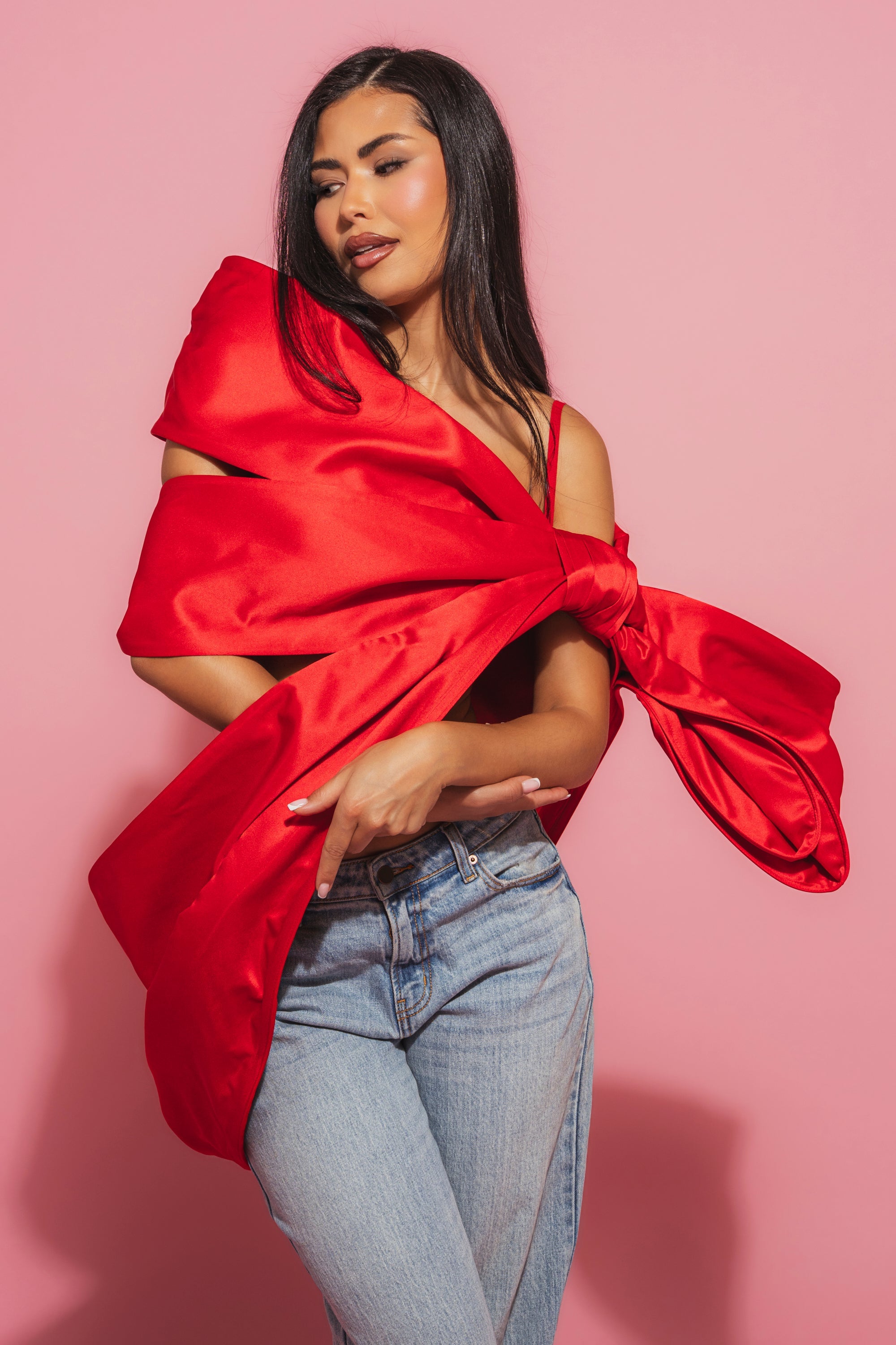 A woman with long dark hair poses against a pink background, wearing the IM THE GIFT BODY ACCESSORY—a bold satin red bow top—paired with light blue jeans. 1