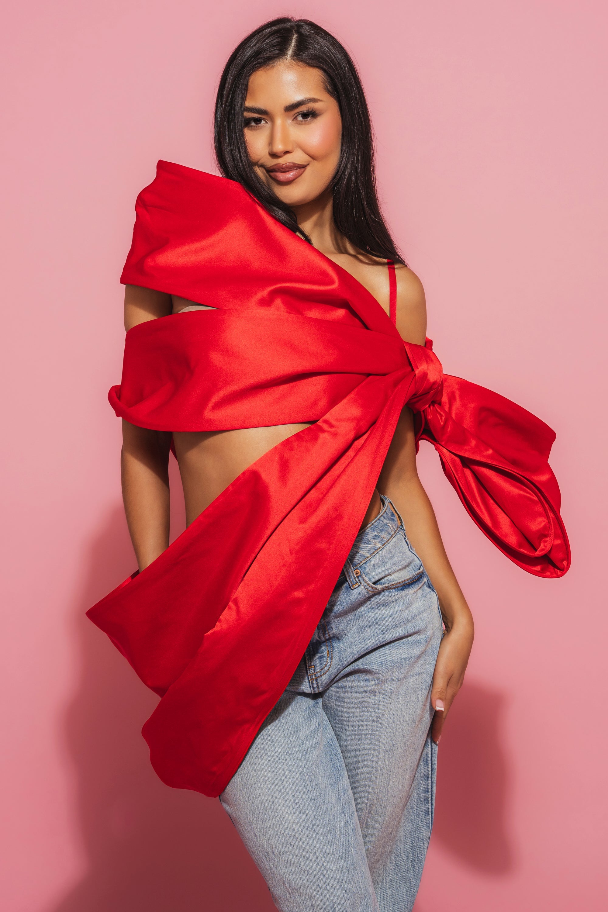 A woman poses against a pink backdrop, wearing blue jeans and the IM THE GIFT BODY ACCESSORY—a bold, asymmetrical red satin piece with an oversized bow draped across her chest and shoulder. 2