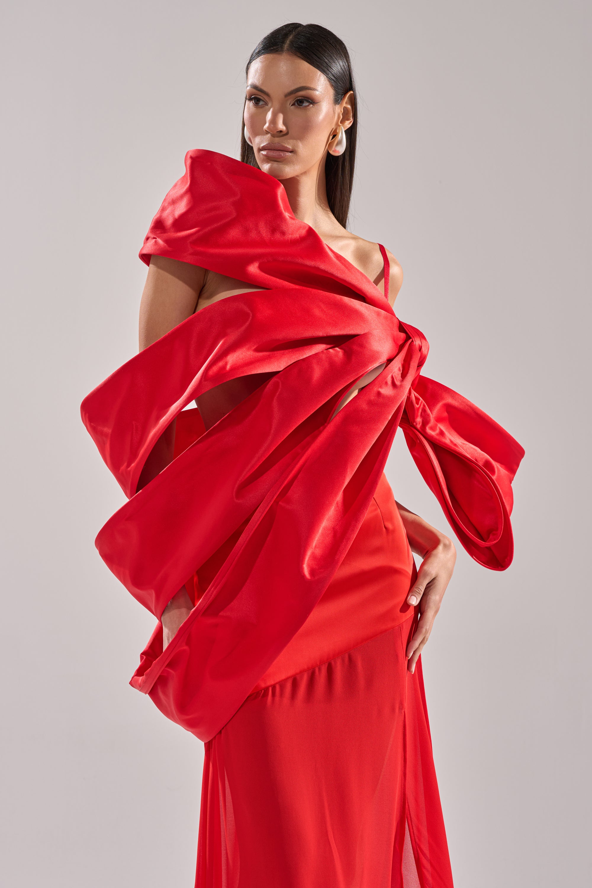 A woman models the IM THE GIFT BODY ACCESSORY, a striking satin red piece with an oversized bow across the chest and shoulder, set against a plain gray background.