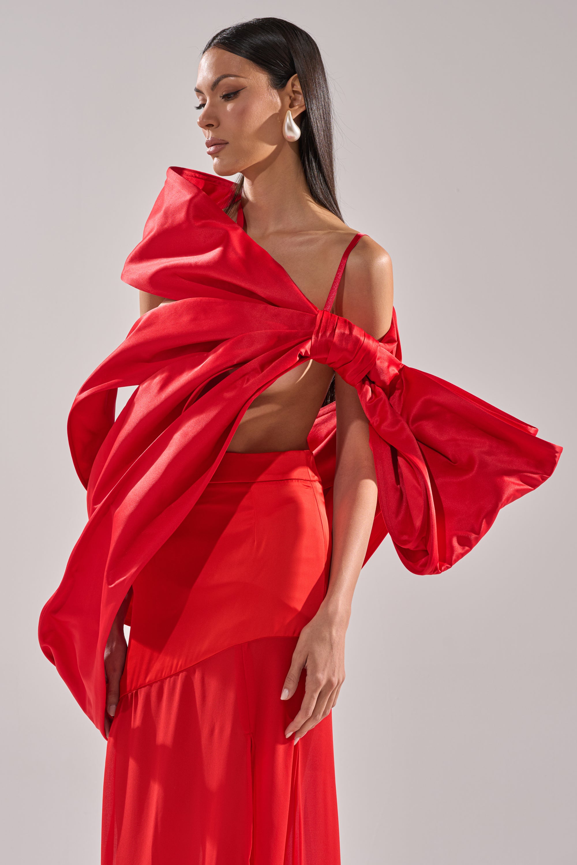 A woman poses in the IM THE GIFT BODY ACCESSORY by AKIRA Label, a vibrant red outfit with a dramatic oversized bow, sleek straight hair, and statement earrings against a plain light gray background.