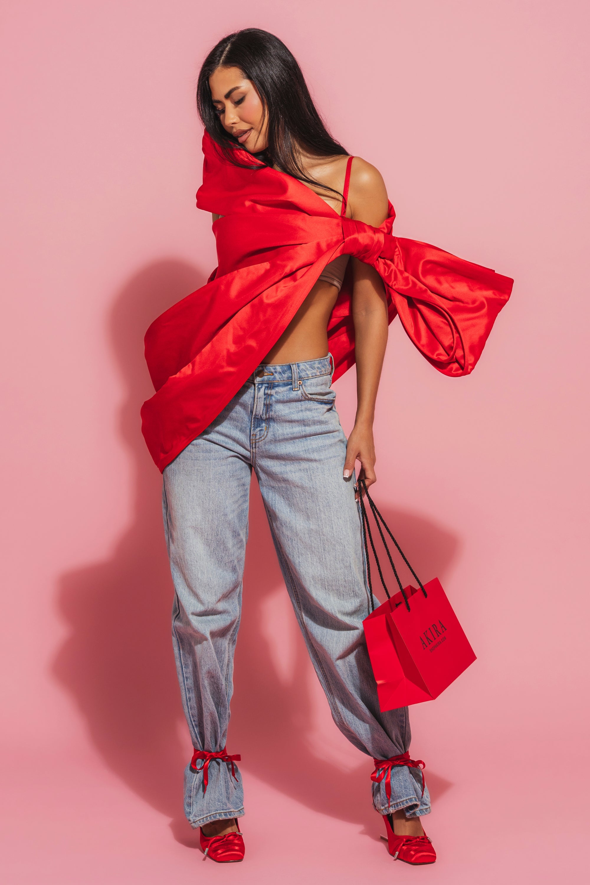 A woman poses against a pink background in a red top, loose light blue jeans, and red ribbon-tied heels, holding a small red shopping bag. She styles her long dark hair with the IM THE GIFT BODY ACCESSORY from AKIRA Label. 3