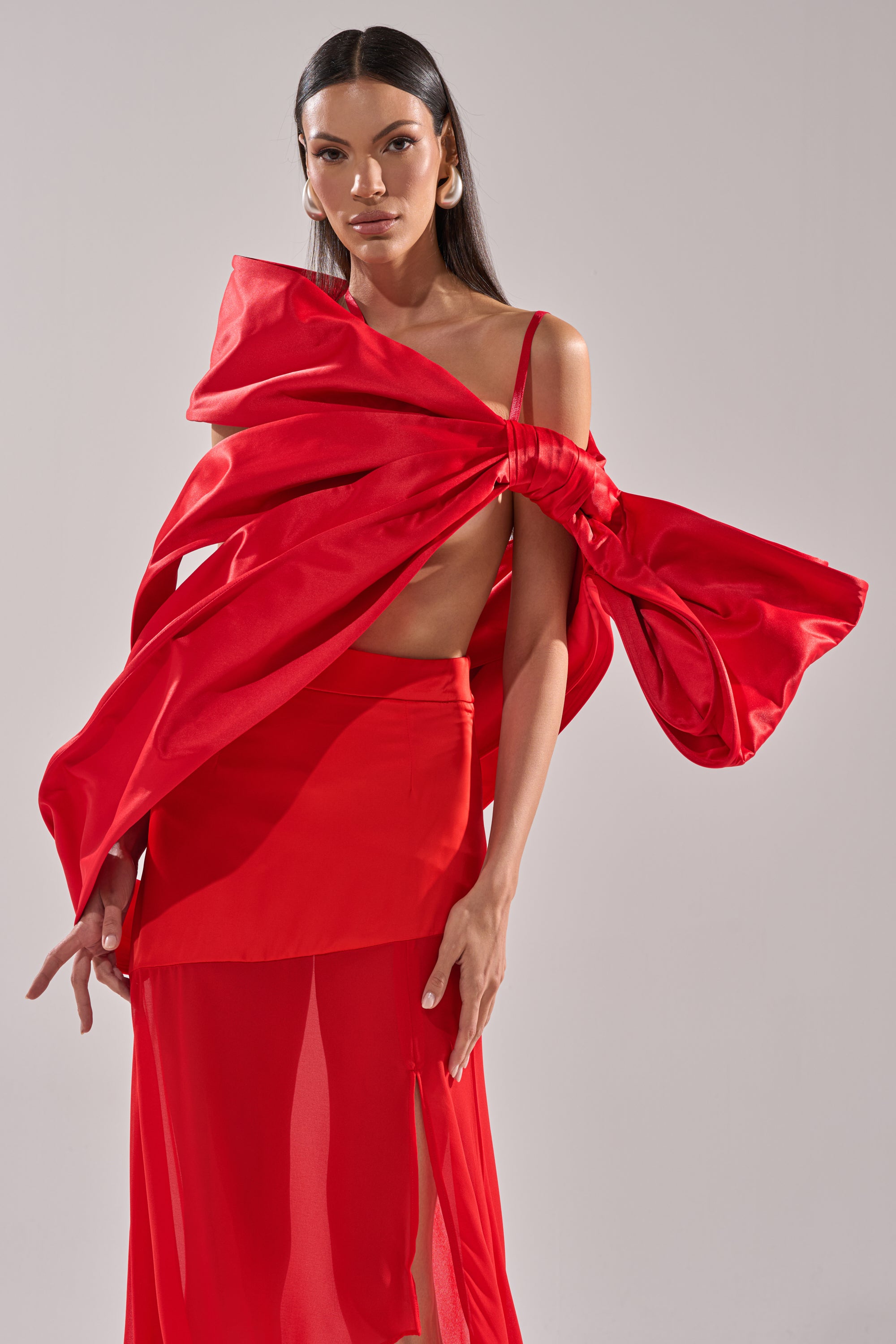 A woman showcases the IM THE GIFT BODY ACCESSORY, featuring a bold red satin oversized bow top and sheer skirt, as she poses confidently against a plain light background.