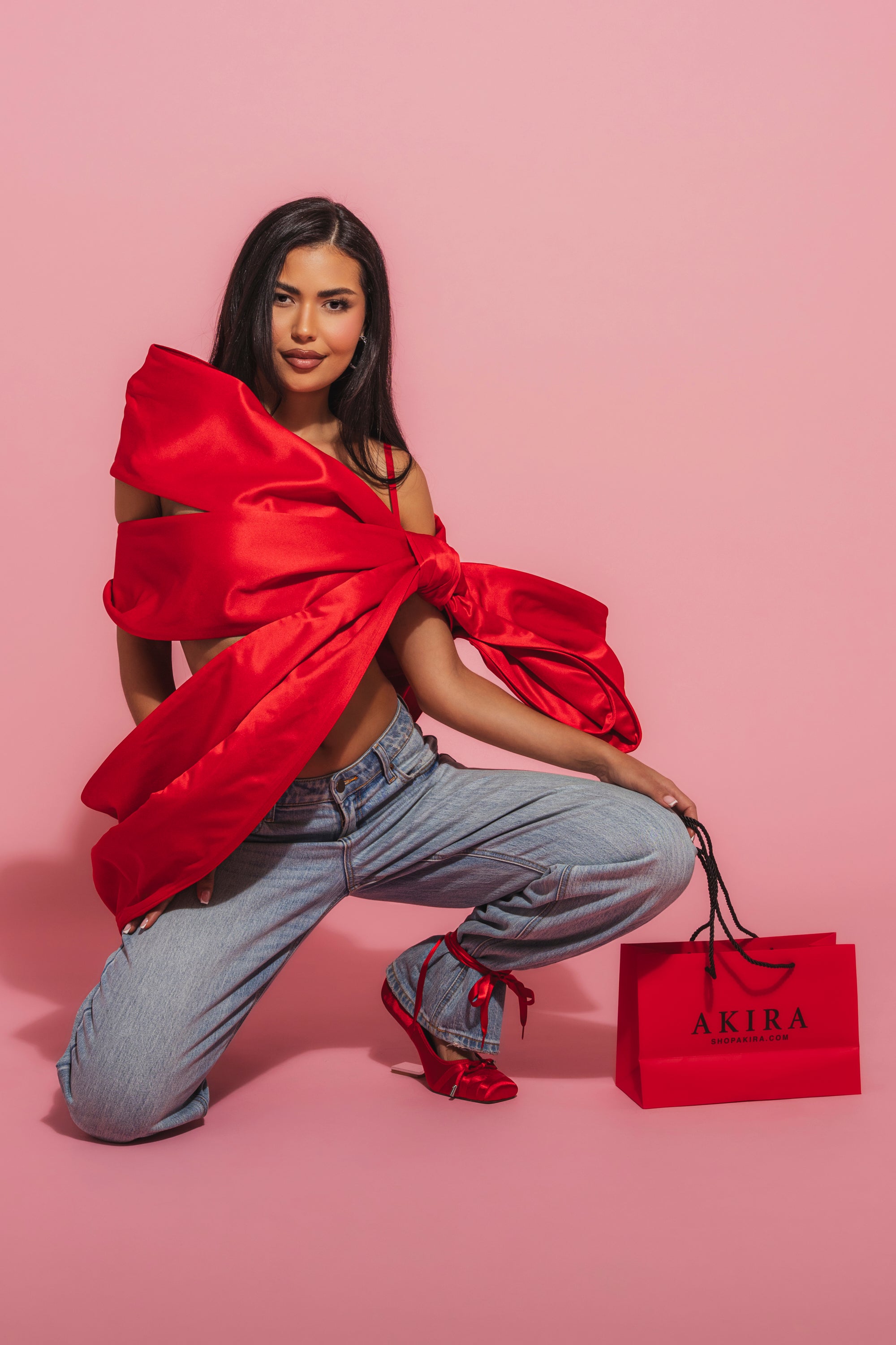 A woman kneels on one knee against a pink background, wearing the IM THE GIFT BODY ACCESSORY, light blue jeans, and red heels. She holds a red AKIRA shopping bag and smiles at the camera. 4