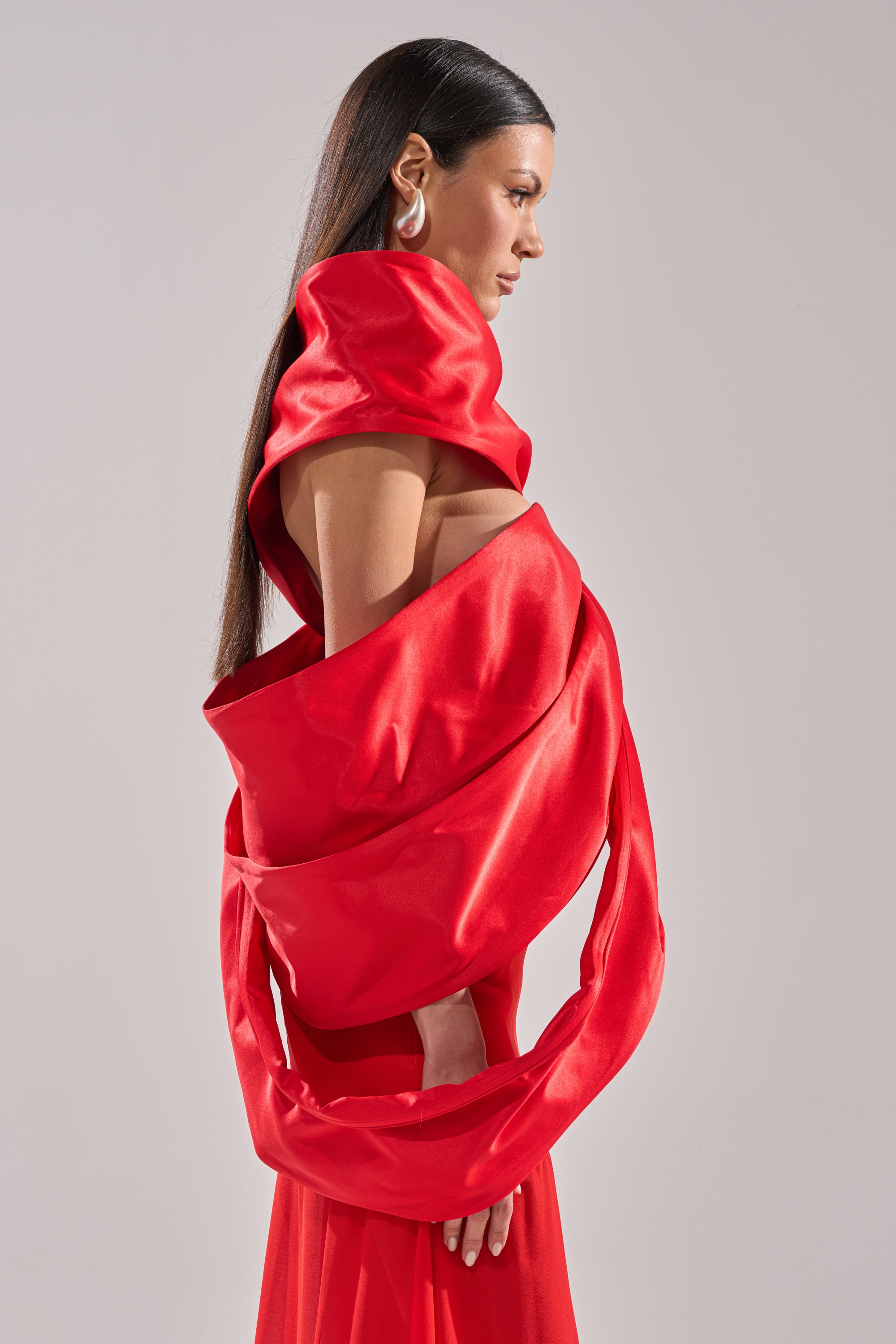 A woman with long straight hair poses in profile wearing the IM THE GIFT BODY ACCESSORY, a bold bright red satin piece with sculptural folds and an oversized bow, paired with large earrings against a plain light background.