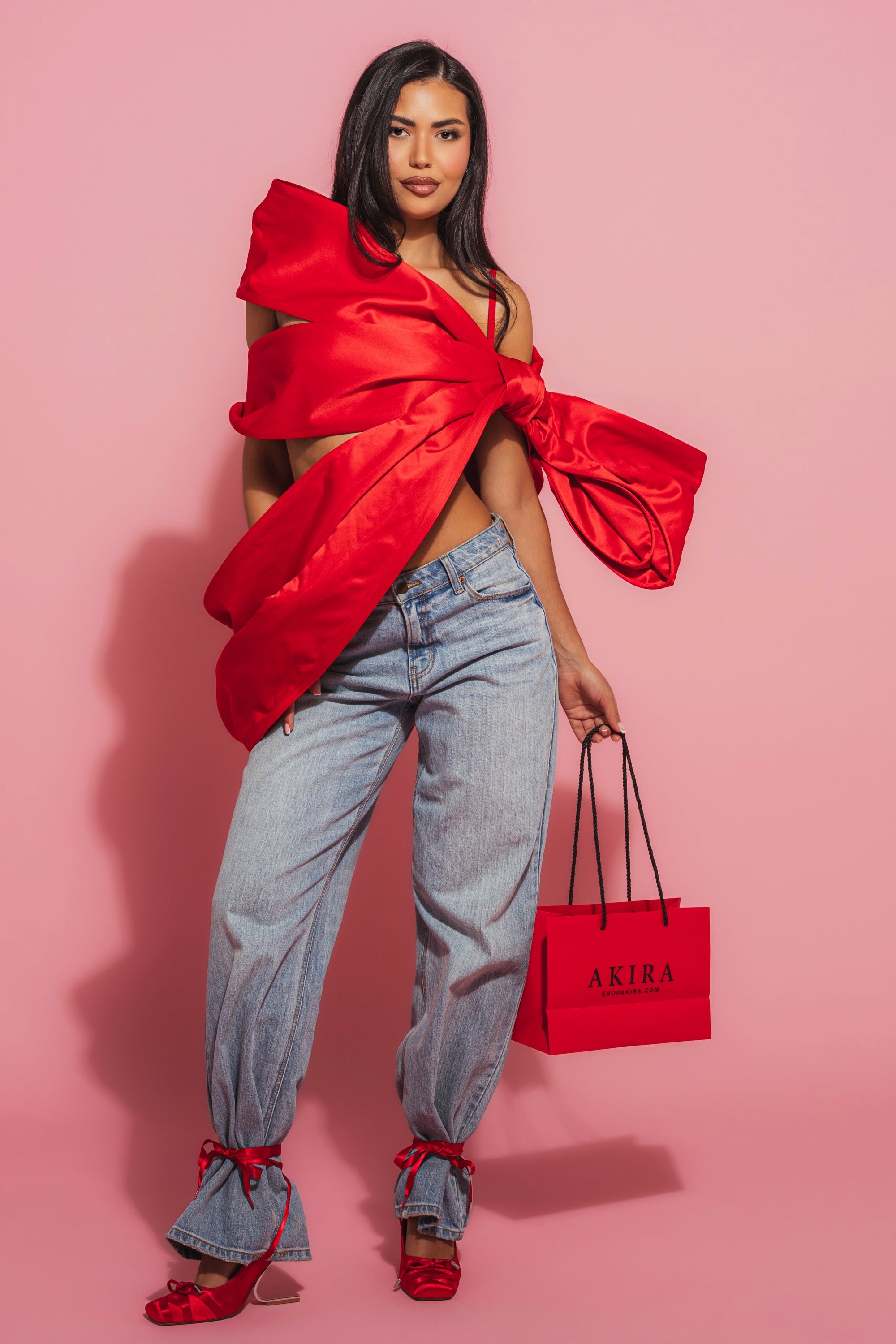 A woman poses against a pink background wearing the IM THE GIFT BODY ACCESSORY as a top, loose blue jeans with red bows at the ankles, red heels, and holding a red AKIRA shopping bag. 5