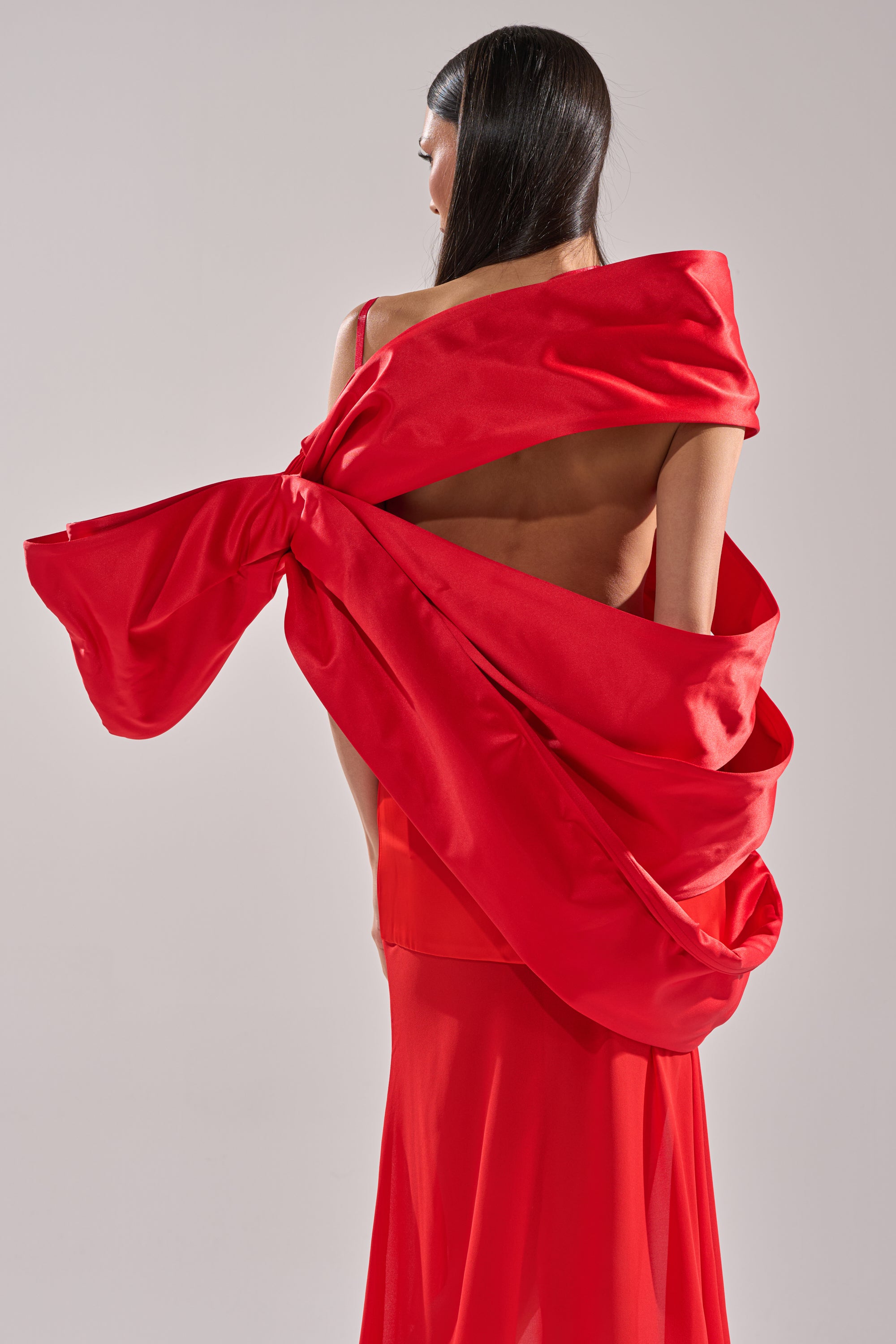 A woman with long dark hair wears the IM THE GIFT BODY ACCESSORY by AKIRA Label—a dramatic red oversized bow that wraps elegantly around her bare back. She stands against a plain light background, facing away from the camera.