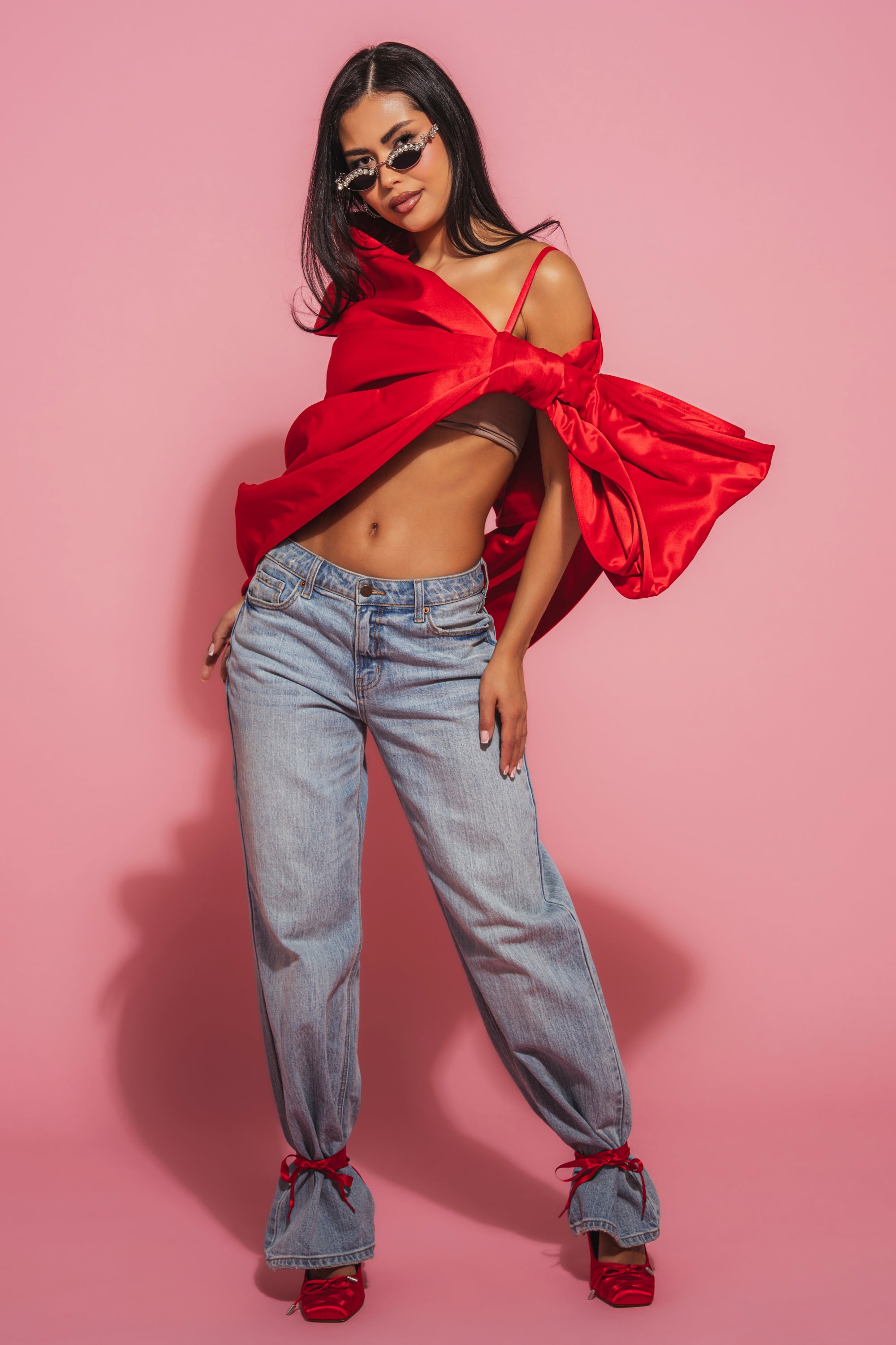 A woman with long dark hair and a confident look poses against a pink background, wearing the IM THE GIFT BODY ACCESSORY—a bright red off-shoulder top with an oversized satin bow—paired with blue jeans and red shoes. 6