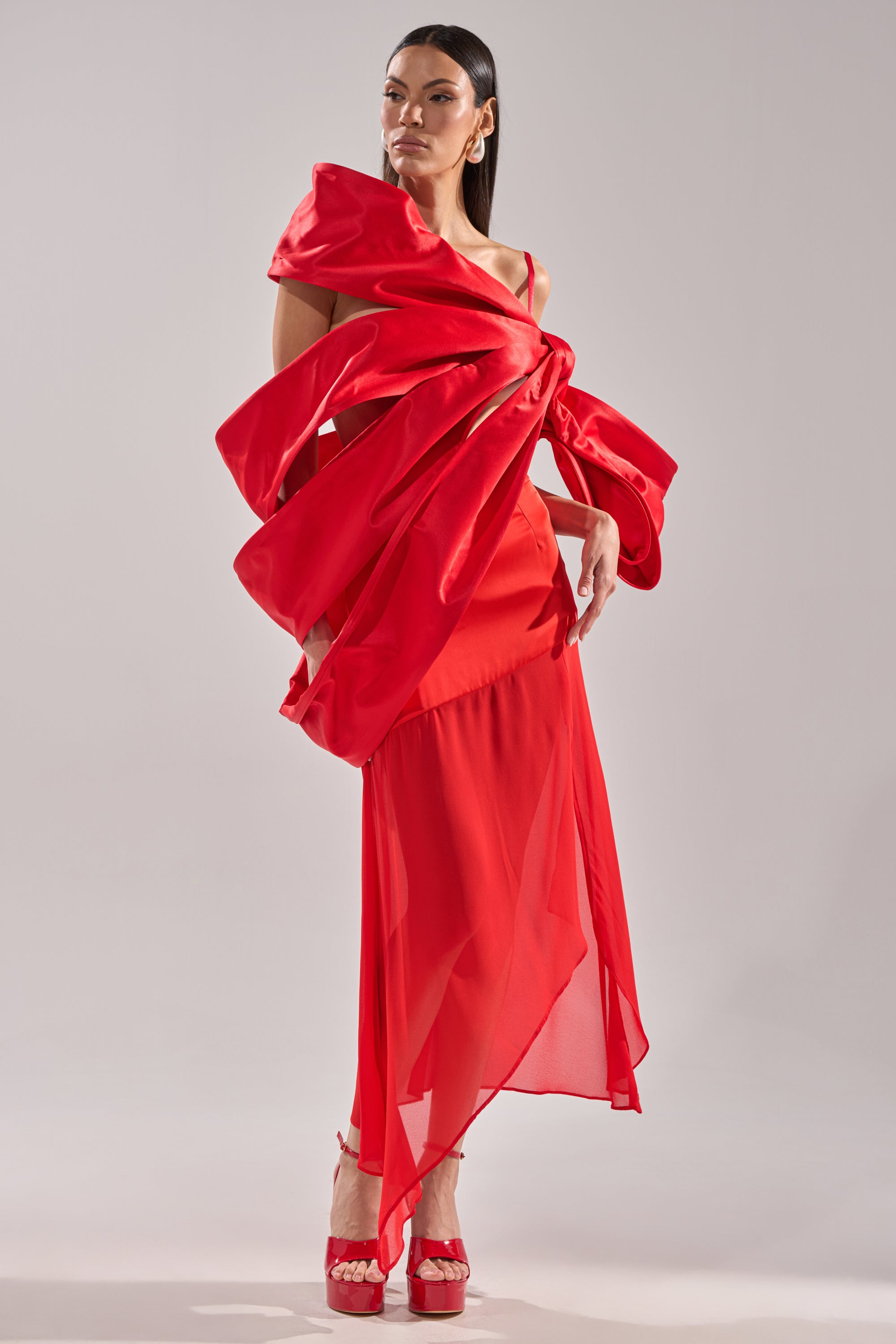 A woman poses in a dramatic, asymmetrical red dress with sculptural ruffles and a sheer skirt, accented by the IM THE GIFT BODY ACCESSORY. She wears red high heels and stands against a plain light gray background.