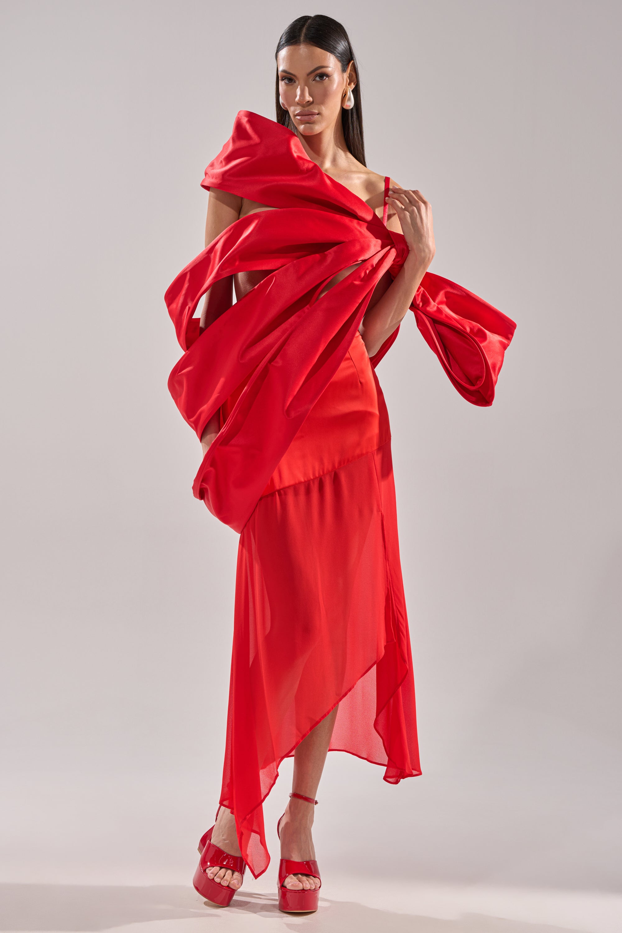 A woman with long dark hair exudes confidence in the IM THE GIFT BODY ACCESSORY: a dramatic red satin dress with a sculptural oversized bow, paired with red open-toe heels and statement earrings against a plain light background.