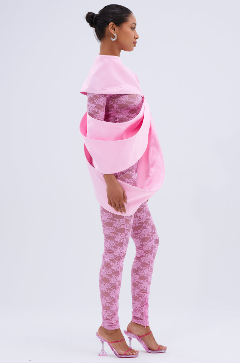 A woman in profile wears a sheer pink lace bodysuit and high heels, styled with the IM THE GIFT BODY ACCESSORY—an oversized satin pink bow draped over her shoulders and waist. Her hair is in a neat bun against a plain light background.