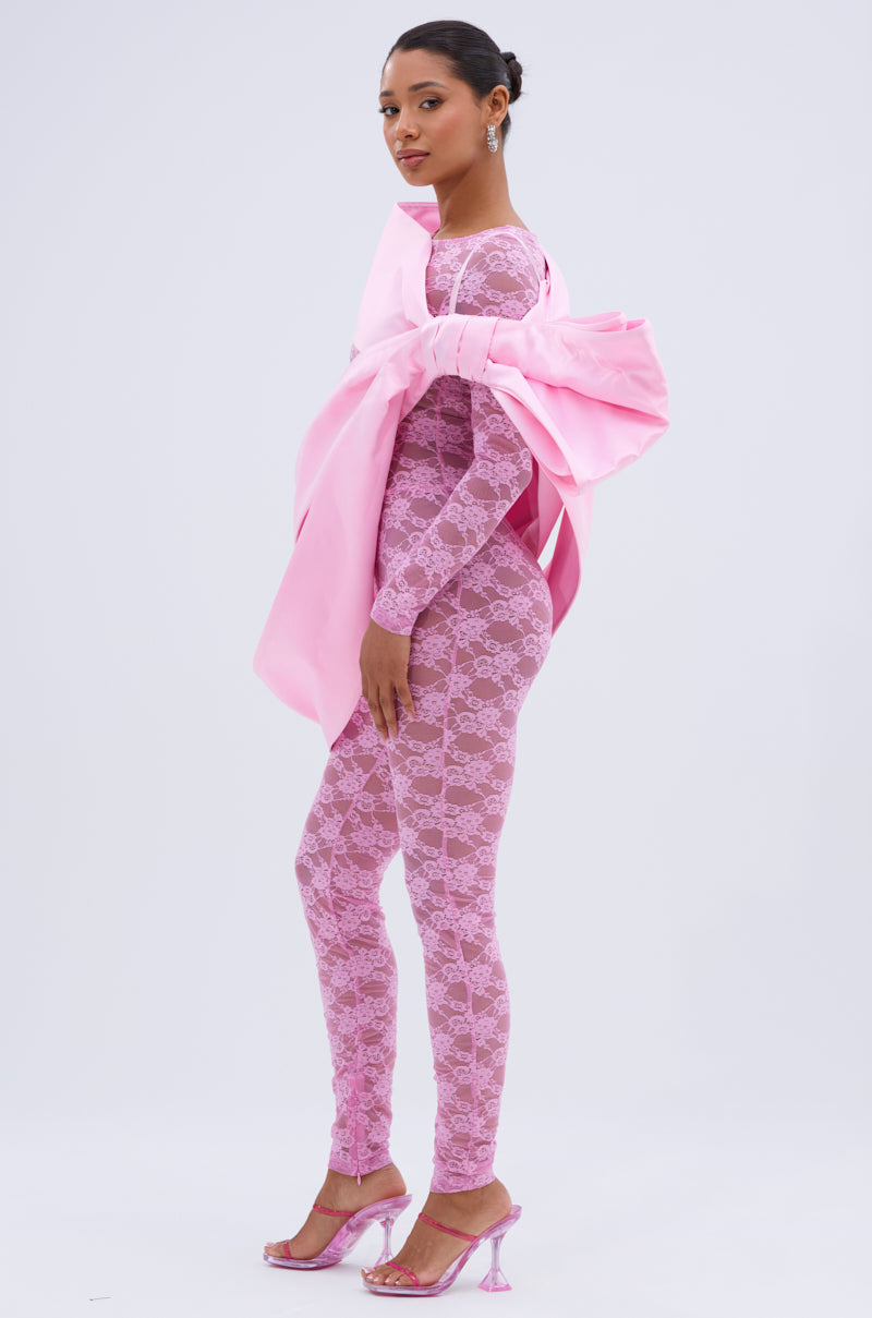 A woman in profile wears the IM THE GIFT BODY ACCESSORY—an oversized pink bow—styled with a fitted, long-sleeved pink lace jumpsuit, pink heels with clear straps, and a sleek bun, set against a plain background.