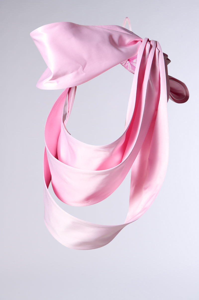 IM THE GIFT BODY ACCESSORY features sculptural satin loops and twists, forming an oversized pink bow suspended against a light gray background.
