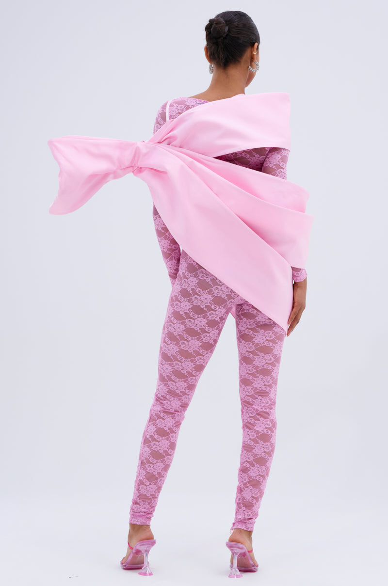 A woman faces away in a fitted pink lace bodysuit and heels, her look accented by the IM THE GIFT BODY ACCESSORY—an oversized pink satin bow draped over her shoulders for a bold, luxe statement against a plain background.
