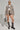 A person poses confidently in the MIMI DENIM TRUCKER JACKET, styled with shorts, a white top, and gray suede boots finished with studs. They complete the look with sunglasses and carry a gray handbag against a plain backdrop.