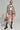 A person poses confidently in the MIMI DENIM TRUCKER JACKET with shorts, paired with a white top. They wear sunglasses, large hoop earrings, grey heeled boots with studs, and hold a grey handbag. The plain background highlights the stylish outfit.