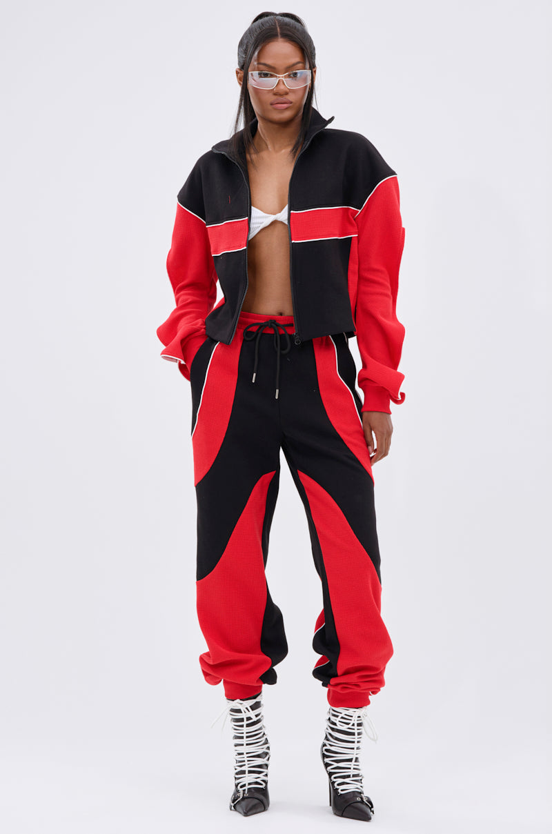 A woman poses in a sporty, modern outfit featuring the LET IT HAPPEN JOGGER, a cropped black and red zip-up jacket, silver bralette, white lace-up heels, and clear futuristic sunglasses.
