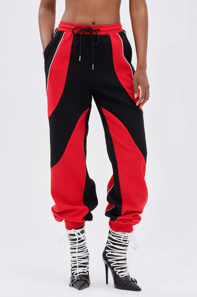 Wearing the LET IT HAPPEN JOGGER, featuring bold red and black colorblocking, paired with white lace-up high-heeled boots and standing against a plain light background.