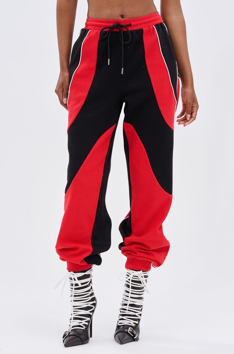 Only the lower body is visible: a person wears the LET IT HAPPEN JOGGER—high-rise, black and red colorblock sweatpants with white piping and black drawstring—paired with pointed black high-heeled boots laced in white.