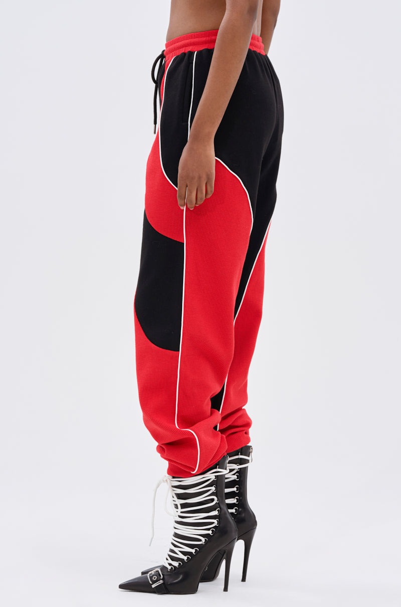 Someone wears the LET IT HAPPEN JOGGER, a high-rise black and red jogger with white piping, paired with black pointed-toe high-heeled boots featuring white lace-up details and buckles.
