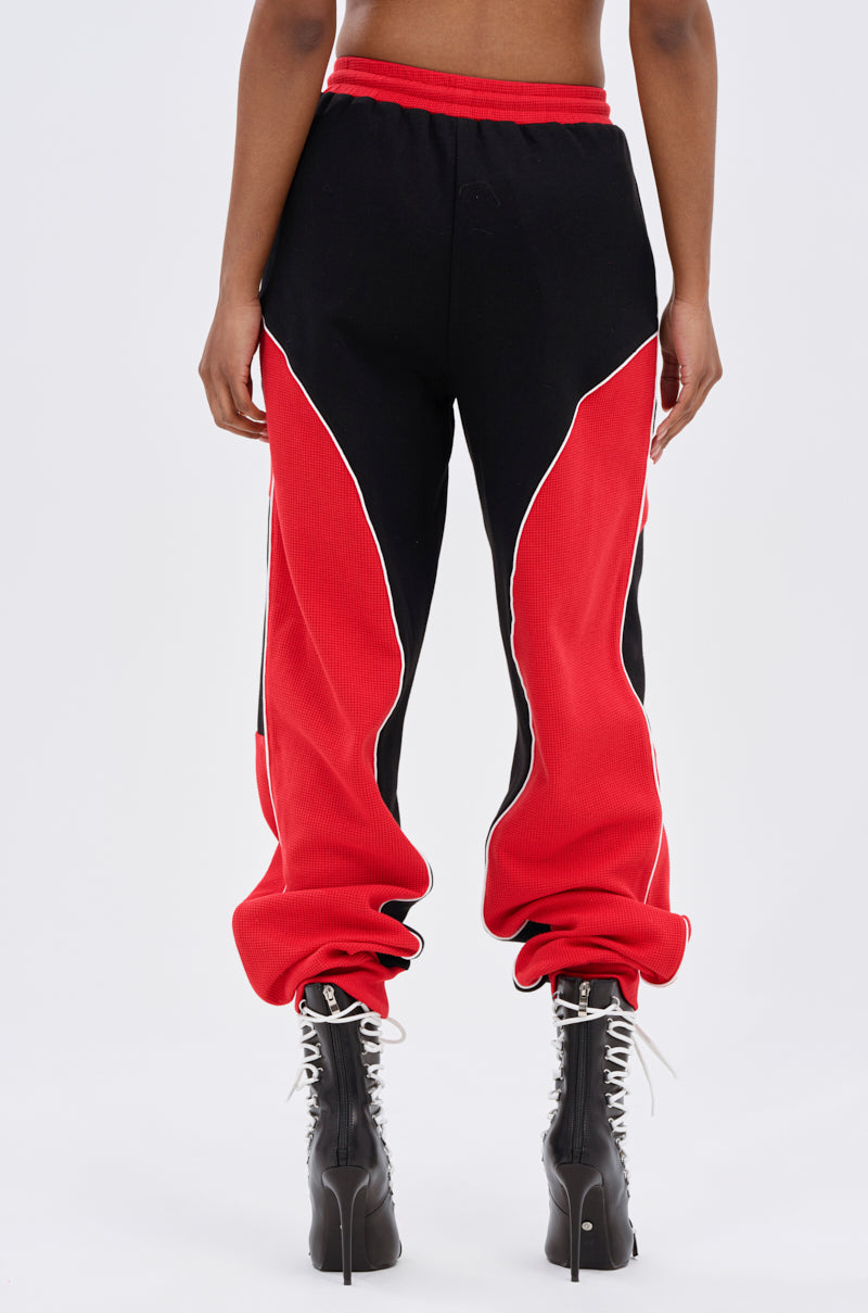A person faces away, wearing the LET IT HAPPEN JOGGER—high-rise jogger pants in black and red colorblocking with white piping, relaxed fit, gathered cuffs—and black high-heeled lace-up boots.