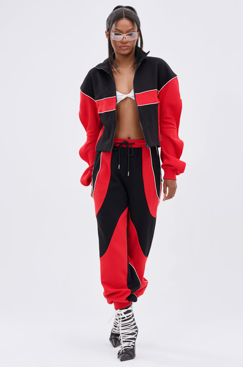 A woman confidently walks against a plain background wearing silver sunglasses, a red and black colorblock track jacket, the LET IT HAPPEN JOGGER with black wave patterns, a silver bralette, and lace-up black-and-white boots.