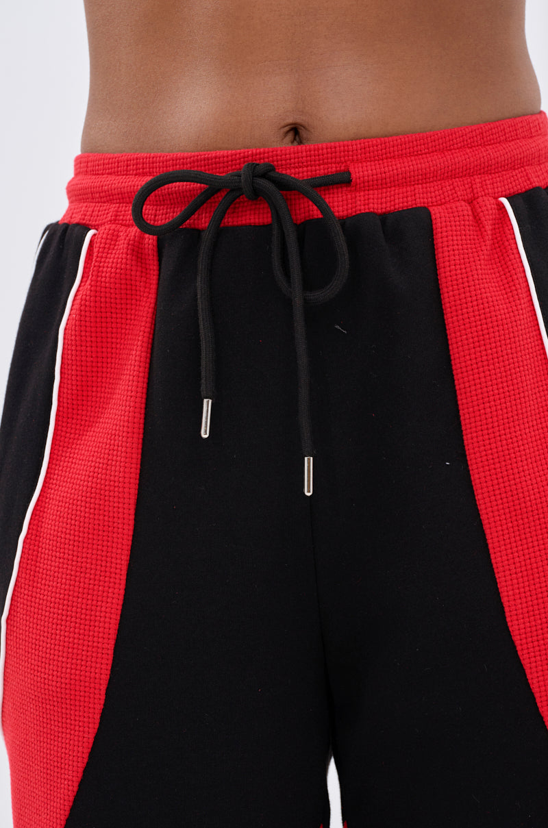 Close-up of a person wearing the LET IT HAPPEN JOGGER—high-rise black shorts with red textured side panels, white piping, a black drawstring waist, and colorblocking that highlights the midriff and lower torso.