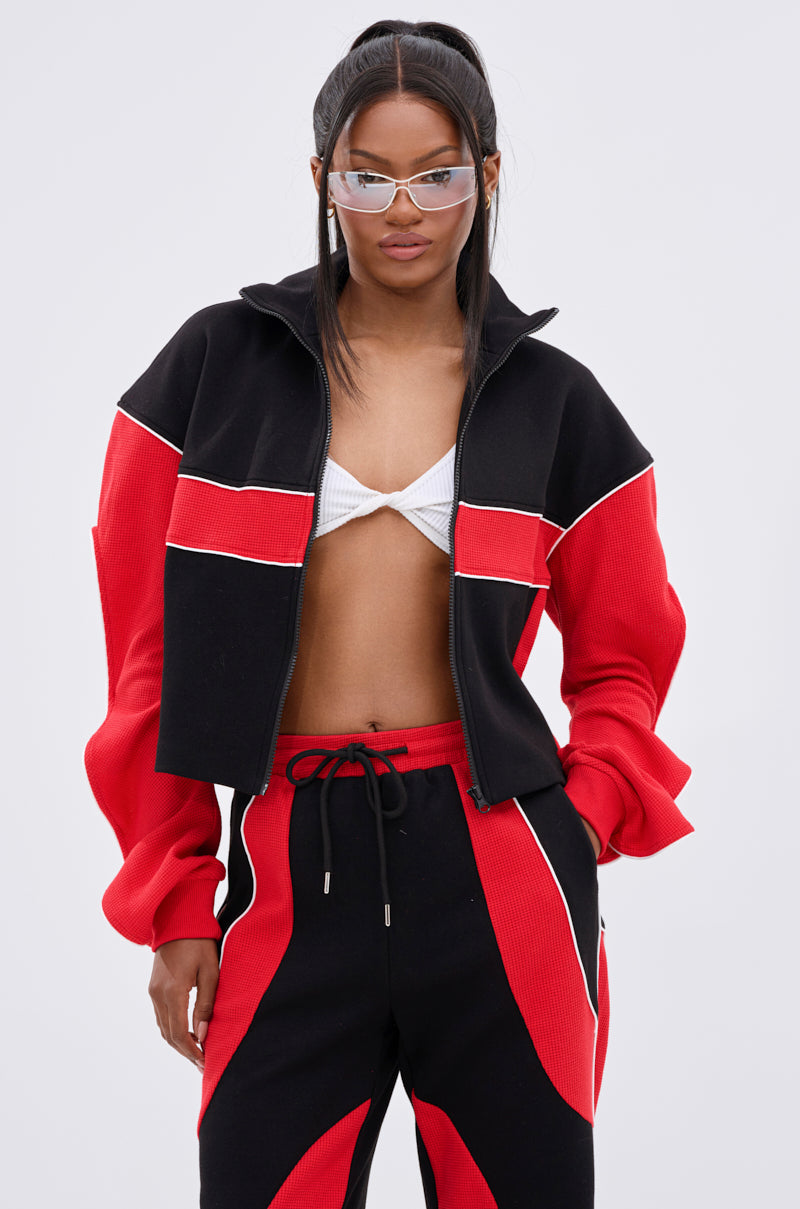 Wearing the LET IT HAPPEN ZIP UP SWEATSHIRT in black and red, matching joggers, a white bra top, and clear futuristic sunglasses, a woman stands confidently with her hands in her pockets against a plain background.