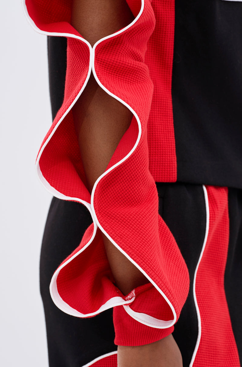 A close-up of a person wearing the LET IT HAPPEN ZIP UP SWEATSHIRT, featuring black and red colorblocking, a textured ruffled red sleeve with white trim, and intricate fabric details.
