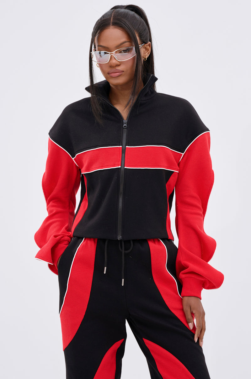 A woman with long dark hair in a ponytail wears clear glasses and a black and red LET IT HAPPEN ZIP UP SWEATSHIRT with matching pants, standing against a plain light background.