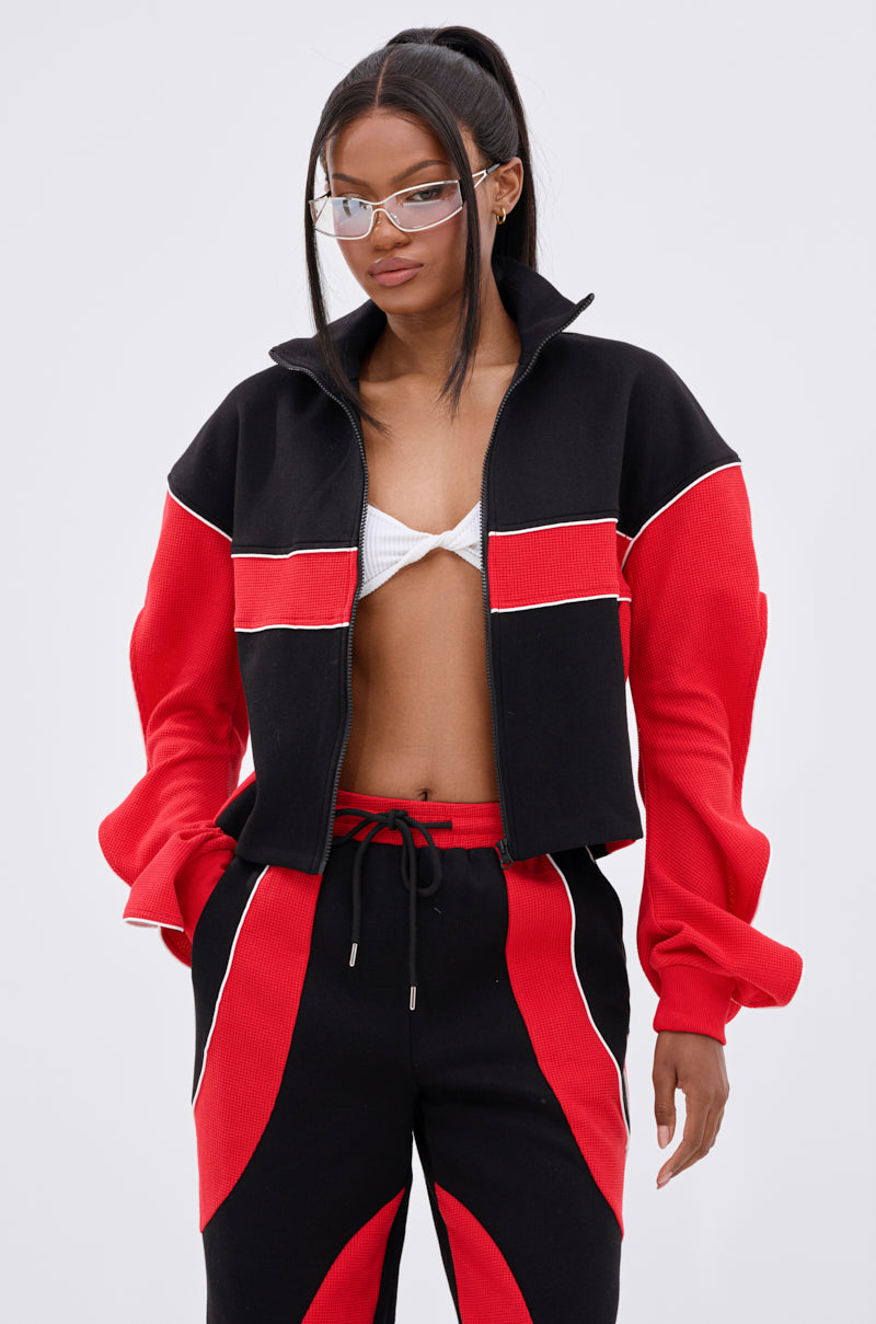 A woman with long straight hair in a high ponytail and clear-framed glasses wears the LET IT HAPPEN ZIP UP SWEATSHIRT over a white bikini top, paired with matching black and red pants.