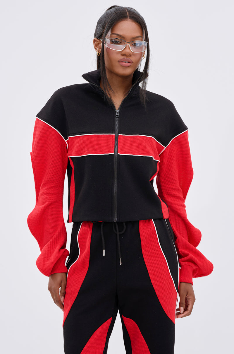 A woman with long dark hair and clear protective glasses wears the LET IT HAPPEN ZIP UP SWEATSHIRT in black and red with matching colorblocked pants and white piping, standing against a plain light background.