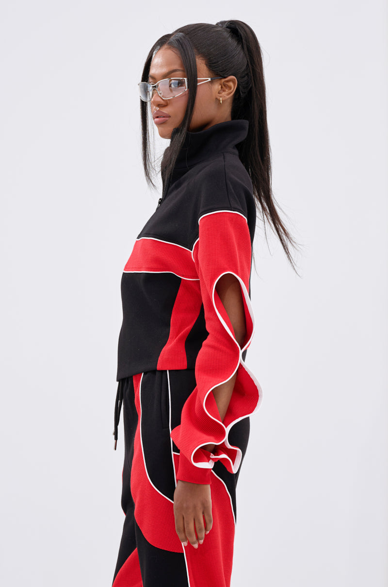 A woman with long, straight black hair in a high ponytail and clear glasses models the LET IT HAPPEN ZIP UP SWEATSHIRT—a stylish black tracksuit with bold red and white colorblocking accents—against a plain light background.
