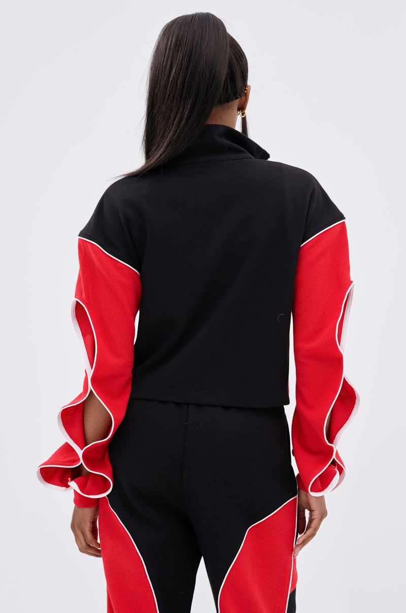 A woman is seen from behind wearing the LET IT HAPPEN ZIP UP SWEATSHIRT, featuring black and red colorblocking with ruffled, white-outlined red sleeves, paired with matching pants. Her long hair is pulled back, showing a partial profile and hoop earrings.